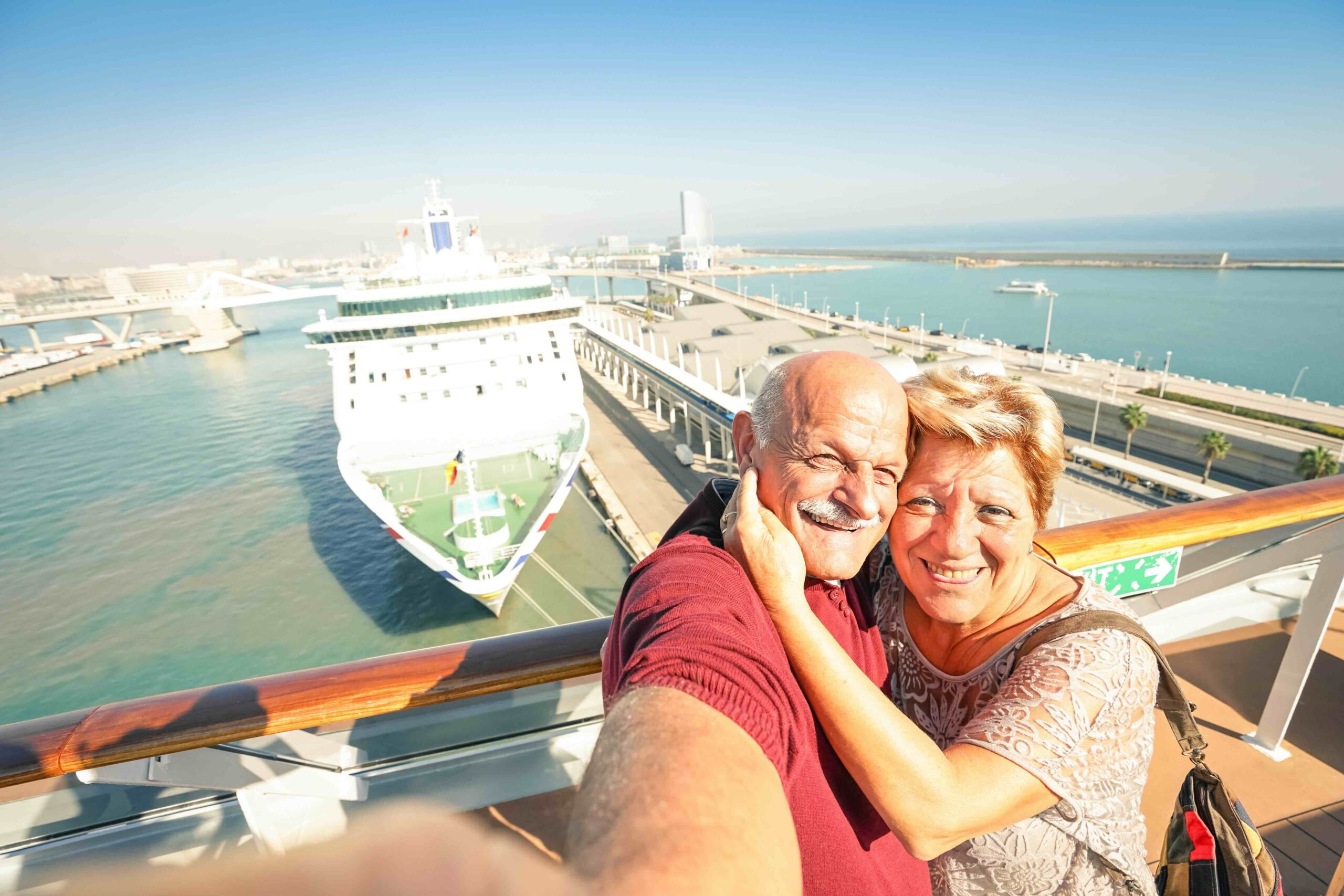 An older couple taking a selfie on the deck of a cruise at port, with a cruise ship in the background. It's easy to go on a cruise on a budget together.