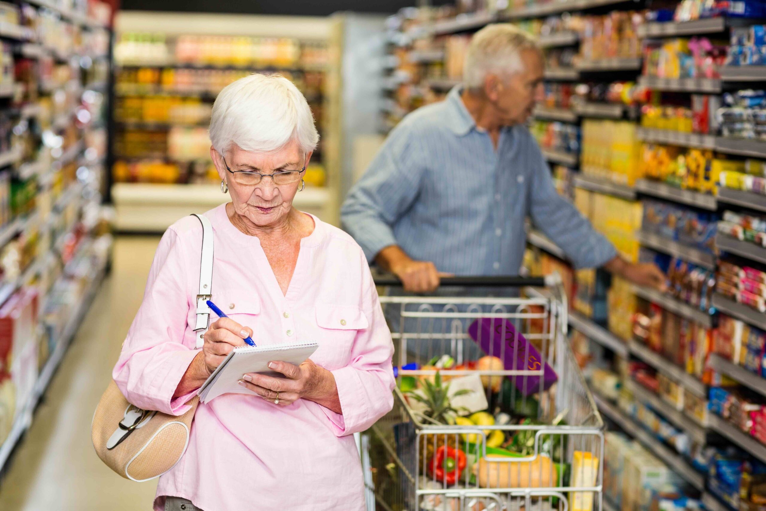 Senior woman with shopping list, her partner pushing the cart. Their cart is full of groceries like produce and shelf stable items. These items can be paid for by the Medicare Advantage grocery allowance if they enroll in a plan that has the benefit.