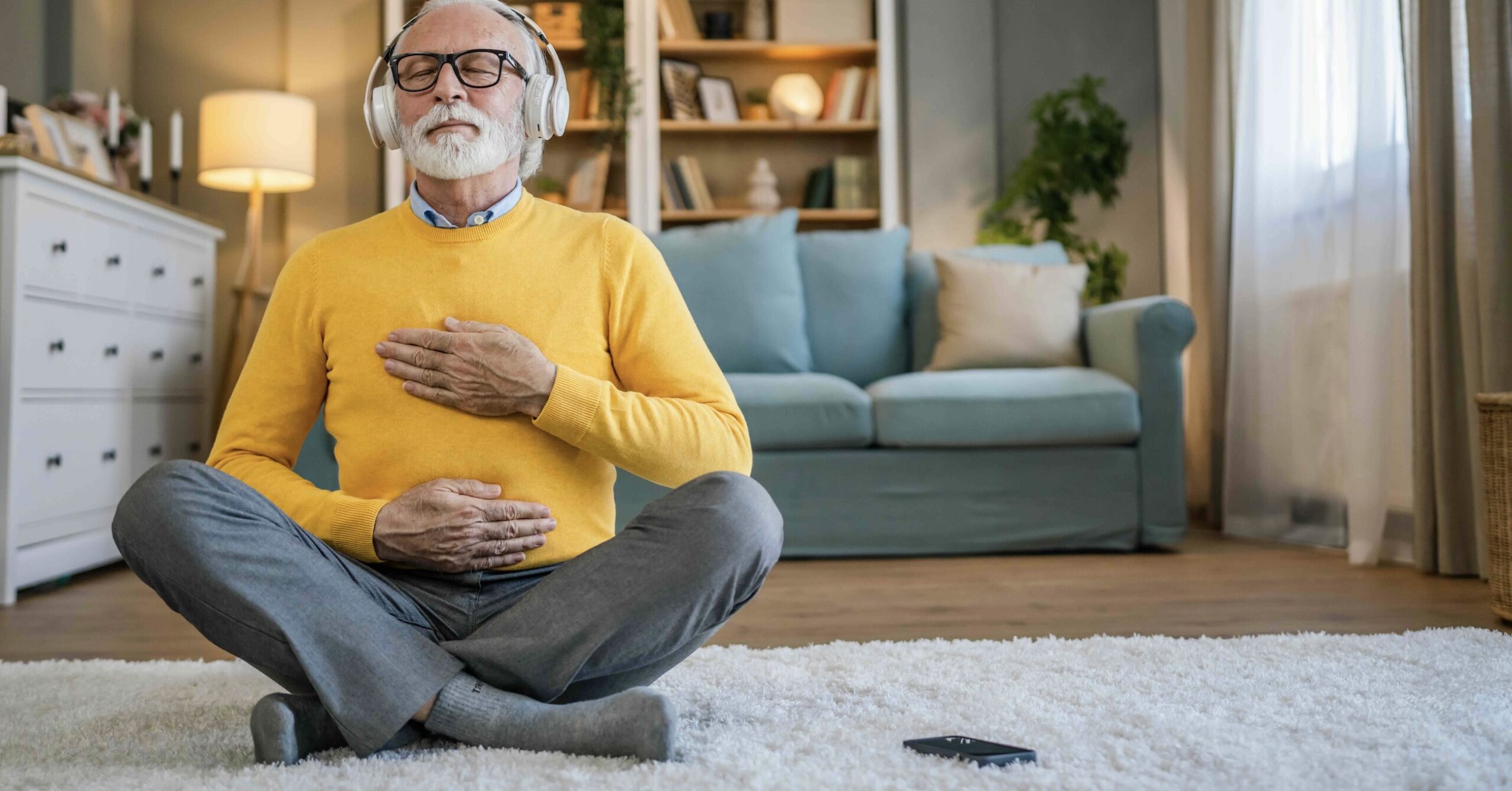 A senior man sitting cross-legged in his living room, wearing headphones and doing a guided meditation through an app or video, keeping his hands on his abdomen to monitor deep breathing. Learn more meditation tips for adults over 60 and get started today.