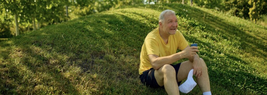 Older man sitting on a hillside holding a cup of water. Wearing light colored clothing and hydrating are two ways to stay cool in the summer.