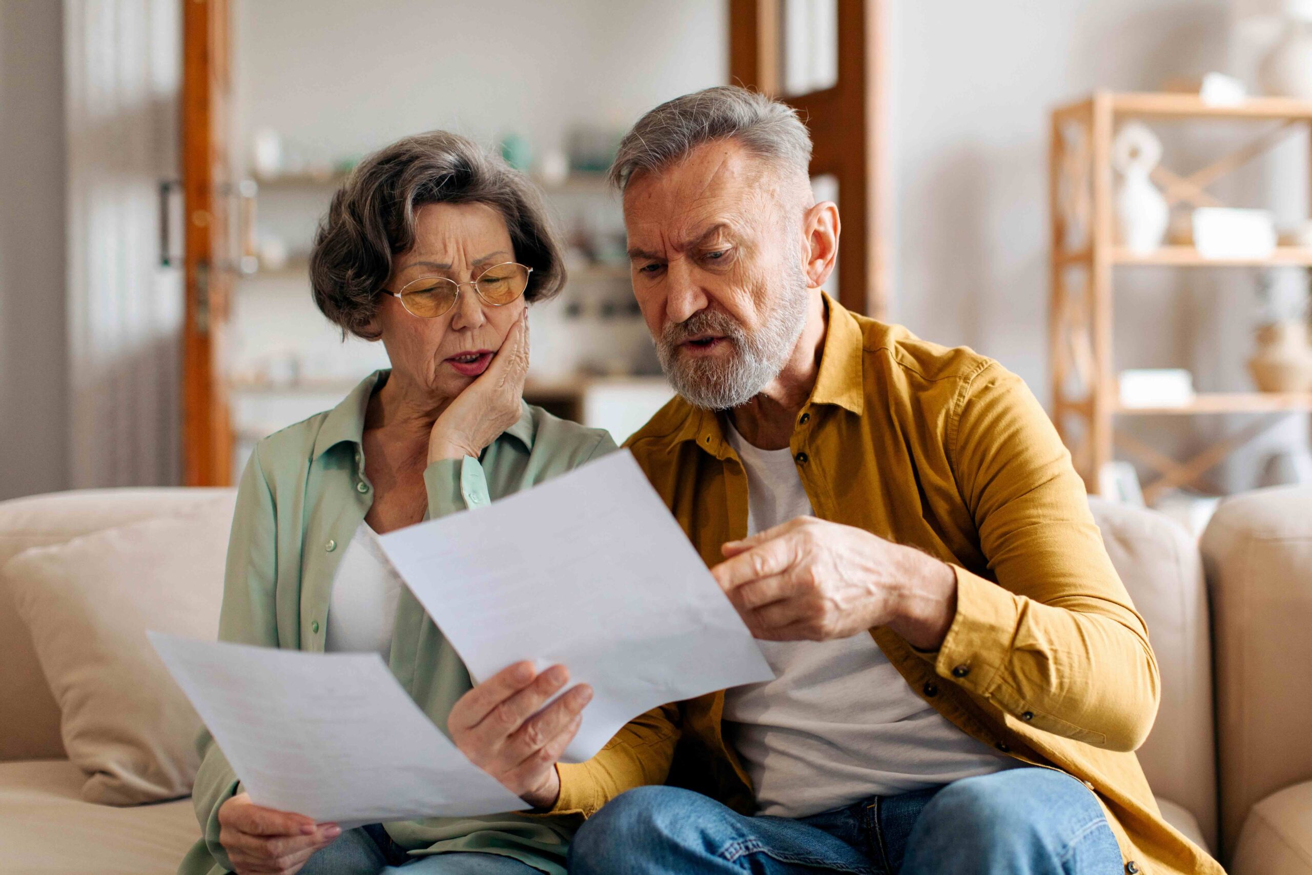 An older man and woman sit on the couch comparing Medicare benefits information. Concept example of a Medicare Advantage plan crosswalk.