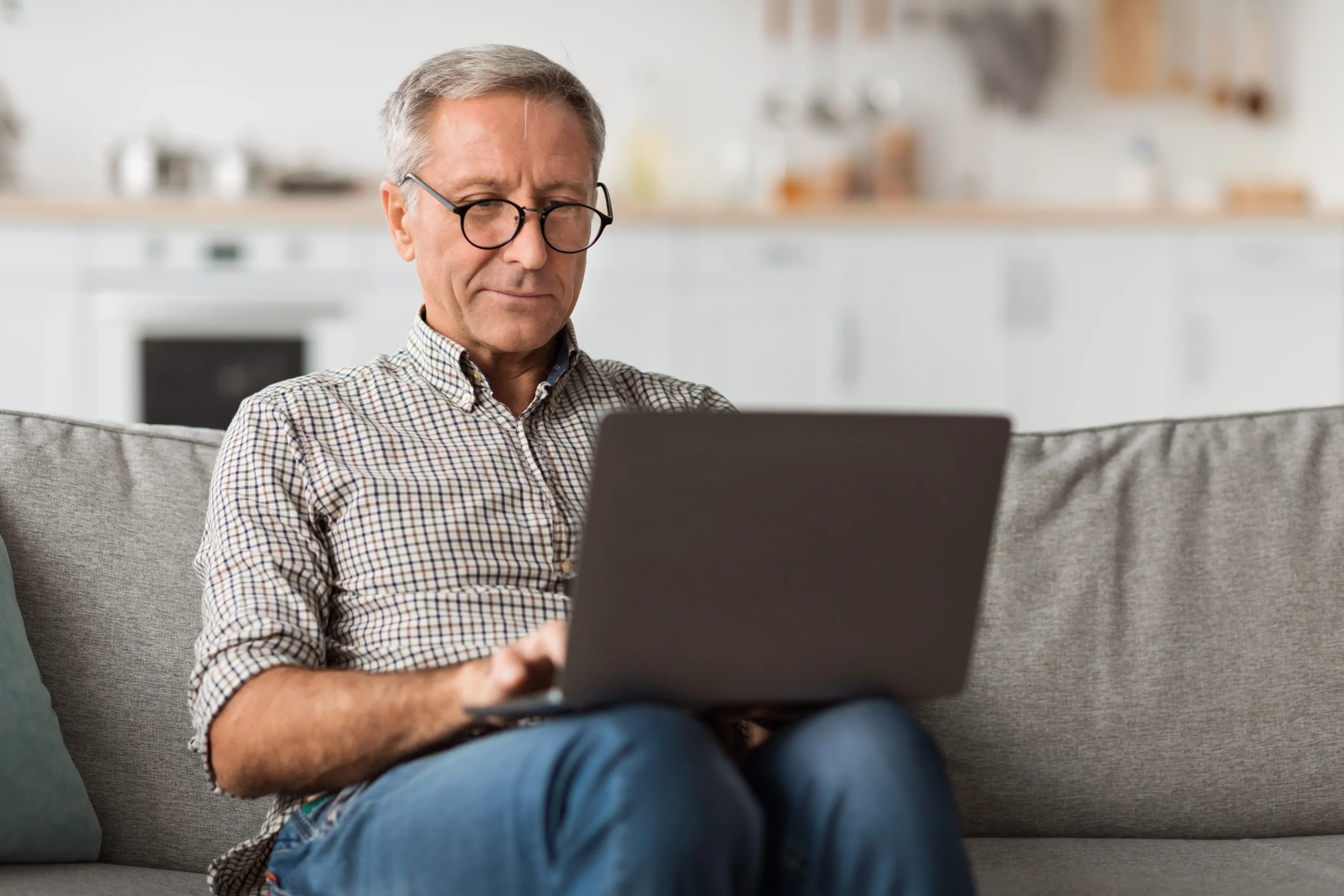 Older man sitting on a sofa typing on a laptop open on his lap. Concept for ACA enrollment on the online marketplace.