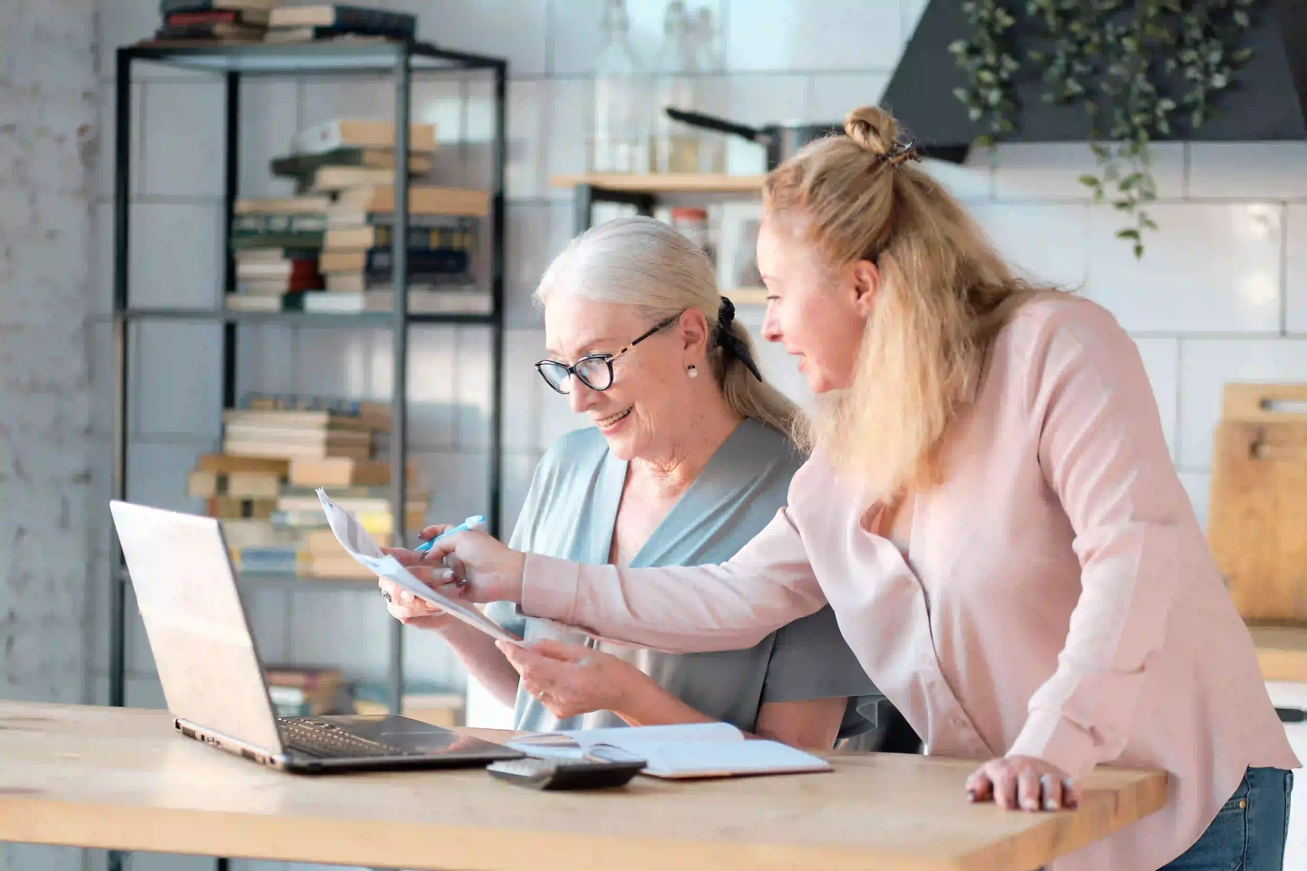 A senior woman and her daughter compare documents at the kitchen table with a laptop.