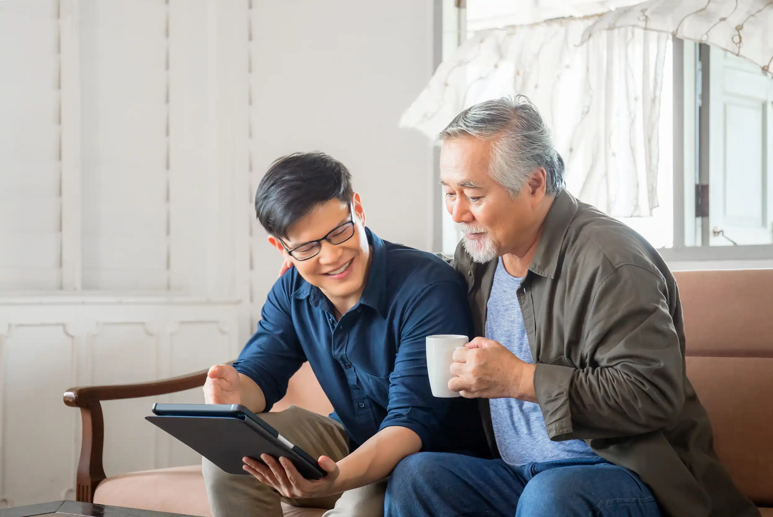 An Asian American man shows his father information on a tablet in the living room. The father holds a coffee cup and has his arm around his son.