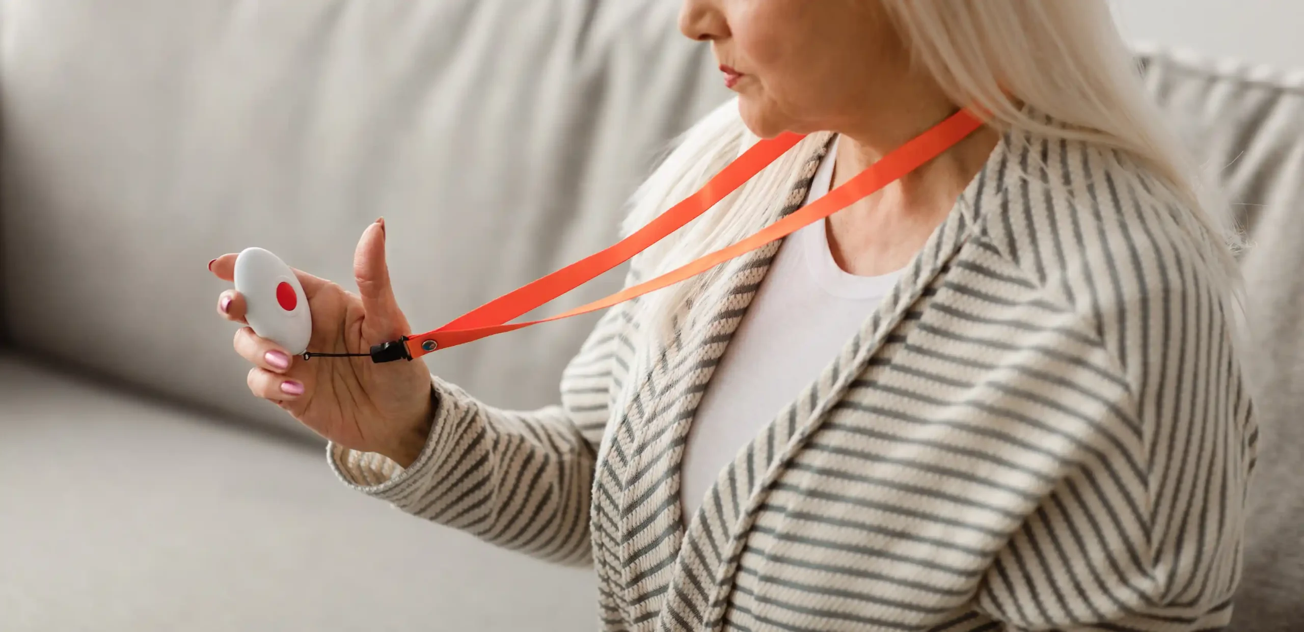 A senior woman sits on the sofa and holds a medical alert system device on a lanyard in her hand.