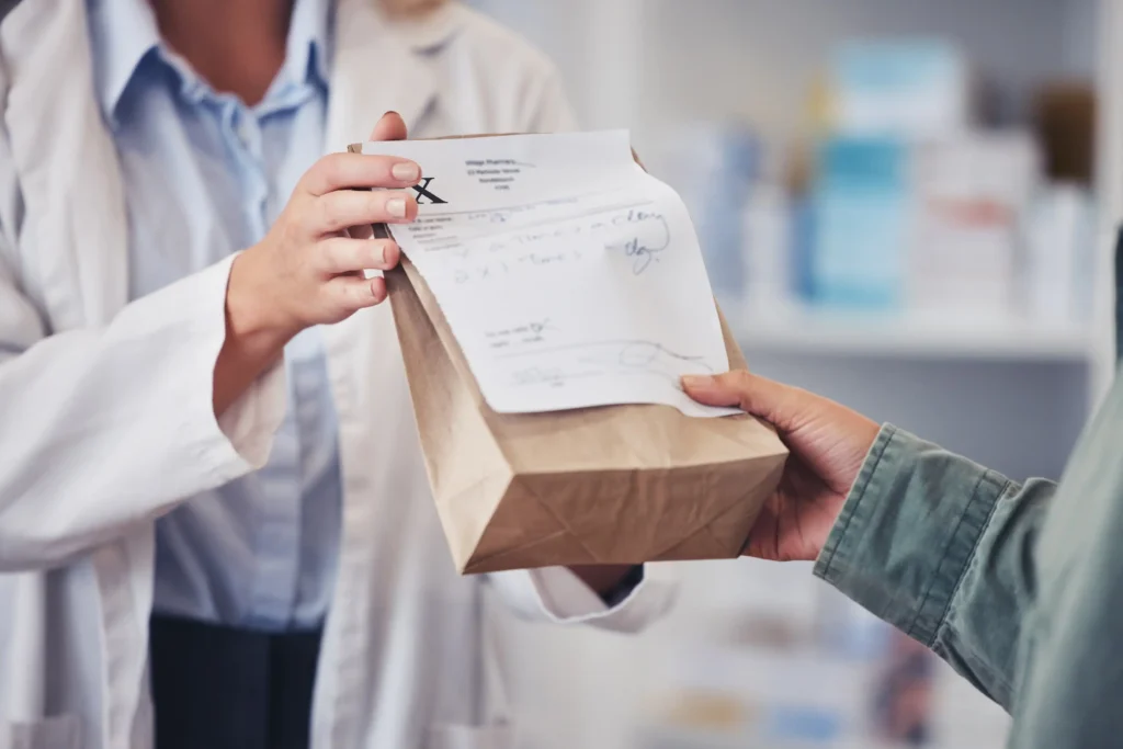 A pharmacist in a white lab coat hands a prescription in a brown paper bag with a receipt stapled to it to a customer, illustrating the process of picking up medications covered under Medicare Part D
