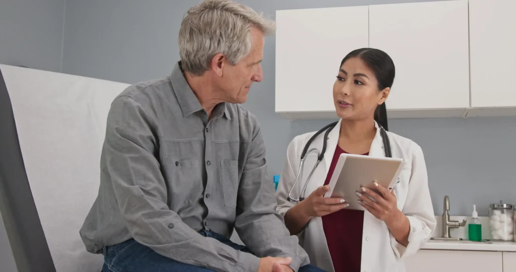 An older male patient consulting with a female doctor wearing a white lab coat and stethoscope. The doctor holds a tablet, appearing to discuss results or options for men's health screenings during a routine medical appointment.