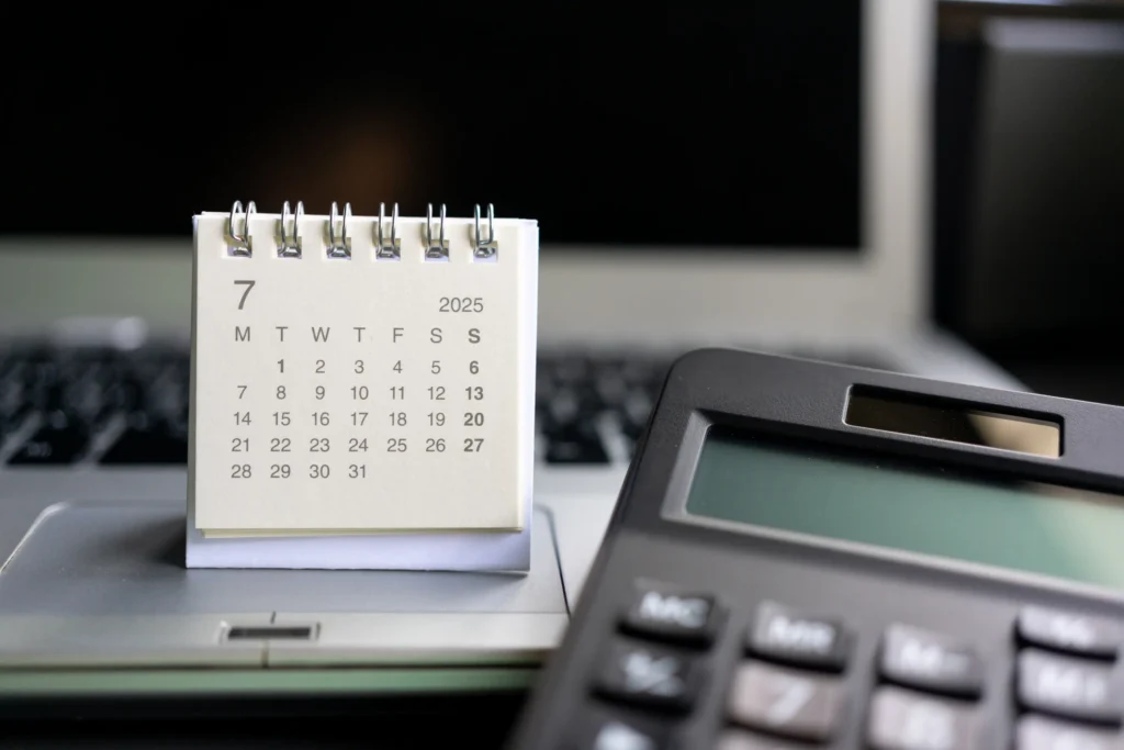Desk calendar open to July 2025 on a laptop with a calculator, representing mid-year Medicare prescription payment plan timelines.