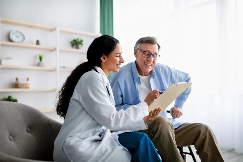 A smiling female doctor in a white coat shows a clipboard to an older male patient in a bright room.