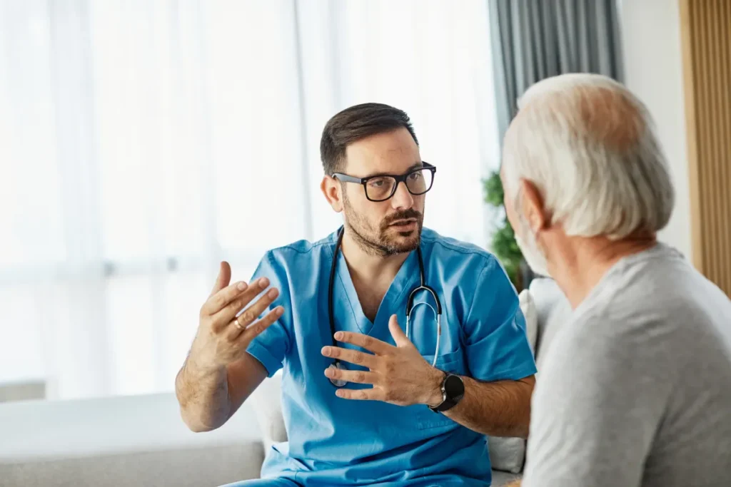 A male doctor in blue scrubs and glasses talks with an elderly patient, gesturing with his hands.