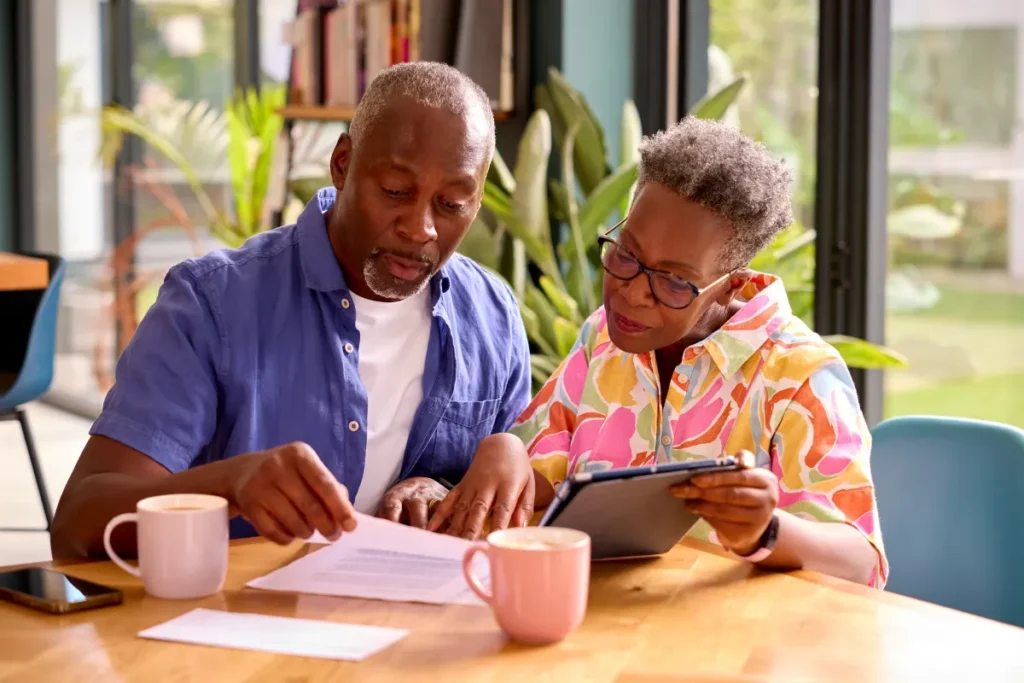 An older Black man and woman comparing HMO vs PPO Medicare Advantage plan documents and a tablet together at a wooden table.