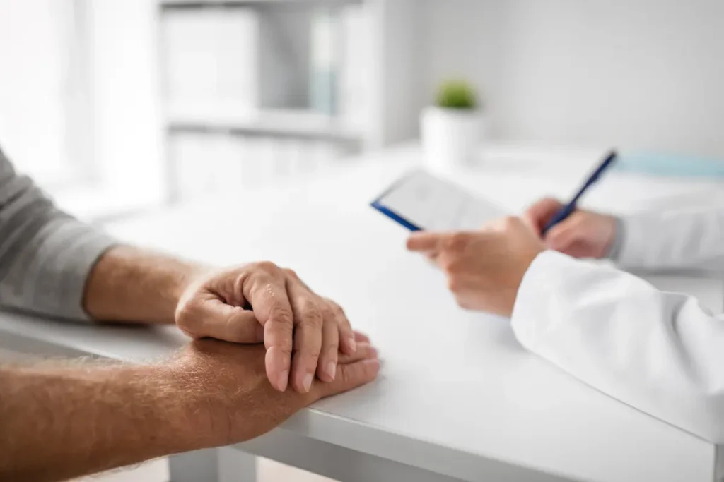 A doctor in a white coat holds a pen while a patient rests their hands on a desk.