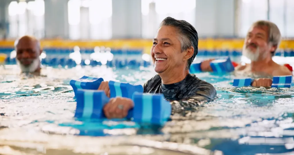 A smiling older woman in a pool holds blue foam dumbbells during a water aerobics class.